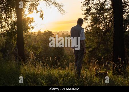 Un homme d'âge moyen marche dans le parc avec son chien sous le ciel doré du coucher du soleil dans la soirée. Promenades en plein air. Famille, animaux de compagnie et concept de voyage. Banque D'Images