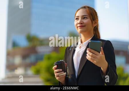 Femme de direction asiatique tenant une tasse de café et utilisant un téléphone mobile dans la rue avec des immeubles de bureaux en arrière-plan à Bangkok, Thaïlande. Banque D'Images