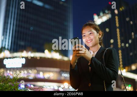 Femme de direction asiatique travaillant à l'aide d'un téléphone mobile dans la rue avec des immeubles de bureaux en arrière-plan la nuit à Bangkok, en Thaïlande. Banque D'Images