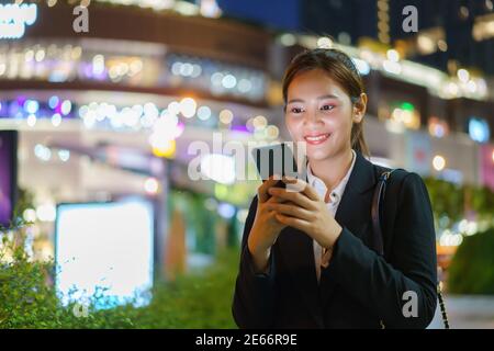 Femme de direction asiatique travaillant à l'aide d'un téléphone mobile dans la rue avec des immeubles de bureaux en arrière-plan la nuit à Bangkok, en Thaïlande. Banque D'Images