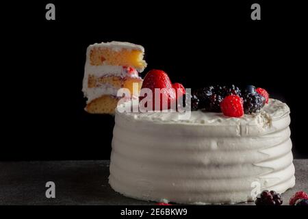 Gros plan d'un gâteau rond avec glaçage blanc crémeux et assortiment de baies utilisées comme garnitures décoratives. Servi sur un fond de granit noir gris foncé Banque D'Images