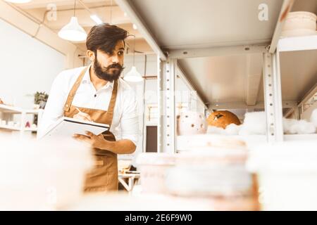 Jeune homme d'affaires séduisant un potter avec une barbe et une moustache travaille dans son atelier. Conserve les enregistrements et transcrites dans un ordinateur portable en inspectant Banque D'Images