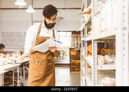 Jeune homme d'affaires séduisant un potter avec une barbe et une moustache travaille dans son atelier. Conserve les enregistrements et transcrites dans un ordinateur portable en inspectant Banque D'Images