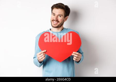 Beau homme européen en chandail disant que je t'aime, petit ami debout avec Saint Valentin coeur rouge, posant sur fond blanc Banque D'Images