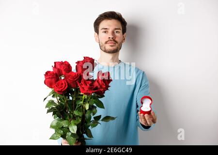 St Valentin et relation. Romantique homme petit ami tenant des roses rouges et montrant l'anneau d'engagement, faisant une proposition le jour des amoureux, debout Banque D'Images