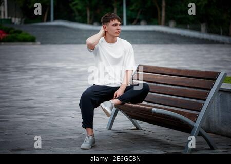 Modèle de t-shirt blanc sur un type assis sur un fond flou d'un carré, sur un banc, regardant dans la distance, vue de face de vêtements. Maquette pour homme Banque D'Images