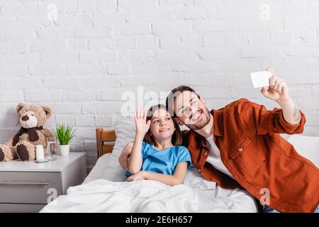 une jeune fille souriante agite la main tandis qu'un père heureux prend le selfie smartphone à l'hôpital Banque D'Images