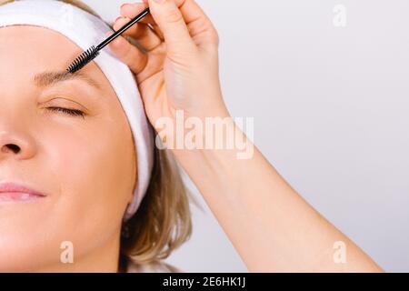 Photo agrandie où la main du maître fait la stratification des sourcils d'une femme avec un bandage sur sa tête. Tendances de la mode sur un blanc Banque D'Images