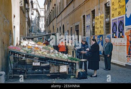 Une cabine de vente de fruits et légumes dans les ruelles de Rome, en Italie, en 1955. Une femme distribue de l'argent pour ses achats. Sur le mur en face se trouvent des affiches, y compris celles pour les montres Certina Swiss et le film "il n'y a pas d'affaires comme le show Business" d'Irving Berlin est un film réalisé en 1954 par Walter Lang. Il a joué Ethel Merman, Dan Dailey, Donald O'Connor, Mitzi Gaynor, Marilyn Monroe, Johnnie Ray et Hugh O'Brian. Banque D'Images