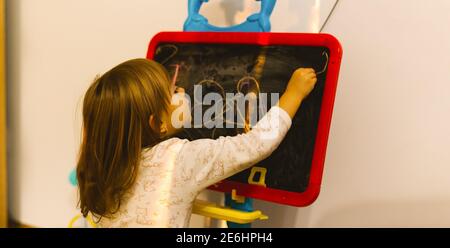 Bannière. La jeune fille d'âge préscolaire tire avec de la craie à la maison sur le tableau noir. Concept de pandémie à domicile. Photo de derrière. Banque D'Images