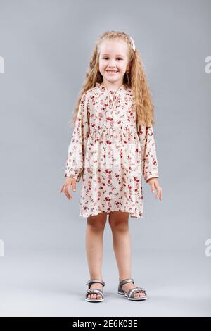 Portrait d'une belle petite fille en forme de curly dans une élégante robe florale tient, rire et regarde dans l'appareil photo dans le studio sur un fond gris Banque D'Images