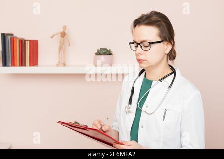 Portrait d'une jeune femme médecin confiante portant un manteau blanc avec stéthoscope tenant le presse-papiers remplissant la forme médicale pendant la visite à domicile Banque D'Images