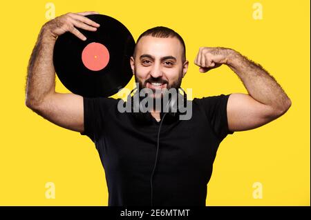 Jeune musicien de dj arabe souriant tenant le disque de vinyle entre les mains et montrant les muscles sur fond jaune isolé Banque D'Images