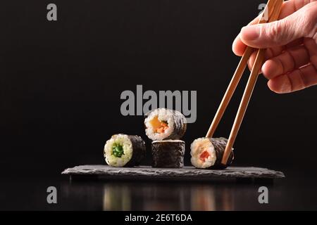 Arrière-plan mains cueillir quatre sushis maki aux légumes assortis avec des baguettes sur plaque de service en ardoise sur table en bois sombre avec fond noir. Avant v Banque D'Images