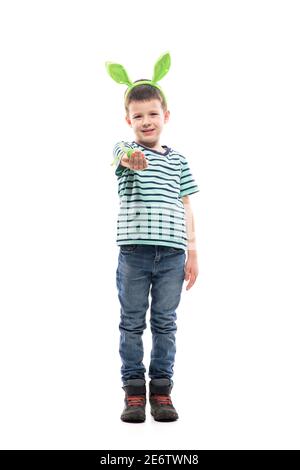 Mignon jeune garçon donnant l'oeuf de Pâques avec le sourire forcé regardant l'appareil photo. Pleine longueur isolée sur fond blanc. Banque D'Images