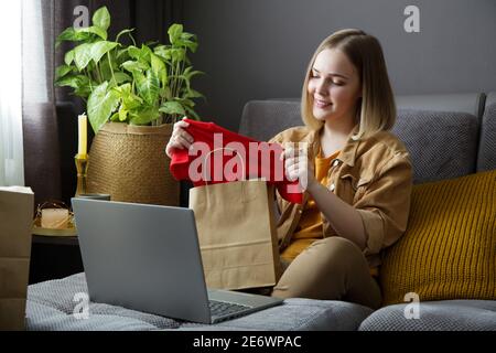 Bonne jeune femme ne déballage commande en ligne de vêtements. Achat en ligne, commande de livraison. Une adolescente se détende sur le canapé en considérant des achats avec Banque D'Images