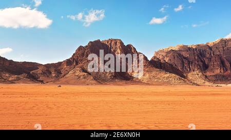 Désert rouge plat avec de grandes montagnes à distance, ciel bleu au-dessus, quelques véhicules tout-terrain en arrière-plan, paysage typique à Wadi Rum, Jordanie Banque D'Images