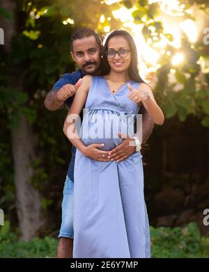 Mère d'être femme tenant son ventre par les deux mains, le père reste derrière et donne le pouce-vers le haut geste, heureux couple posant pour une photo en extérieur soirée tim Banque D'Images