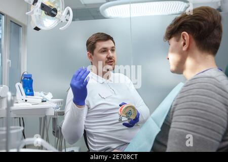 Jeune dentiste de sexe masculin travaillant à sa clinique, parlant à un patient Banque D'Images