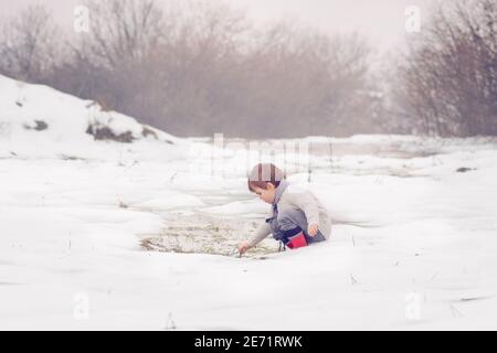 Petit enfant caucasien enveloppé dans une écharpe portant un chandail, jouant dans la neige et le flaque d'hiver. Banque D'Images