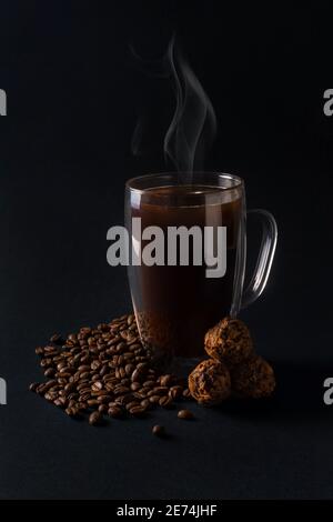 Café noir chaud dans une tasse de verre avec truffe de chocolat et beaucoup de grains de café sur fond sombre. Petit déjeuner café amour concept. Vue avant Banque D'Images