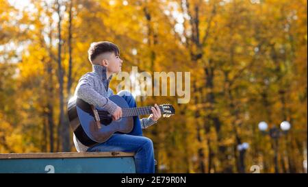 un jeune garçon joue de la guitare acoustique noire le jour de l'automne Dans le Parc Banque D'Images