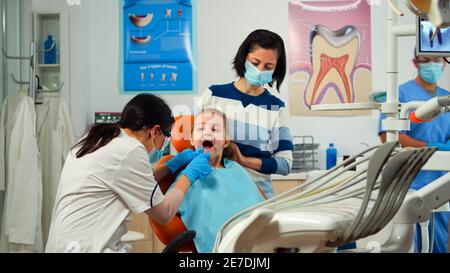Stomatologue médecin vérifiant la santé des dents du jeune patient à l'aide d'outils dentaires travaillant dans un bureau stomatologique moderne avec l'infirmière homme. Mère et fille médecin traitant pour consultation périodique Banque D'Images