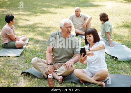 Couple senior assis sur l'herbe et discutant du tracker sportif sur leurs gadgets pendant l'entraînement sportif dans le parc Banque D'Images