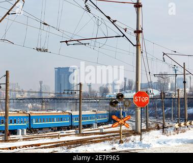 Un panneau d'arrêt rouge à un passage à niveau sur fond de voies ferrées, de voitures de train bleues et de paysage urbain à l'entrée d'une ville d'hiver. Banque D'Images