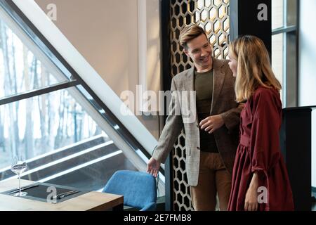 Joyeux jeune homme élégant en tenue décontractée et élégante qui regarde son petite amie blonde tout en l'aidant à s'asseoir sur le velours bleu chaise par table de service Banque D'Images