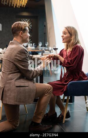 Joyeux jeune homme élégant regardant sa fille surprise pendant placer l'anneau d'engagement sur son doigt par la table servie pendant dîner Banque D'Images