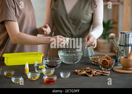 Jeune femme versant du colorant vert liquide dans un bol avec fondu masse de savon tandis qu'une autre femme le mélangeant avant de faire cosmétique produits Banque D'Images