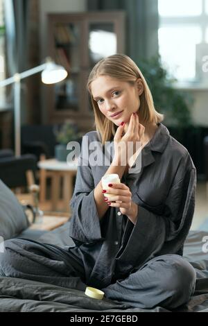 Jeune blonde jolie femme en pyjama gris application naturelle à la main produit cosmétique sur le visage tout en étant assis sur le lit le matin Banque D'Images
