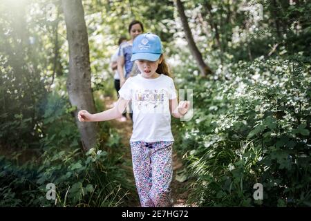 Jeune fille à cheveux longs en t-shirt et casquette de baseball marche à travers la forêt boisée au soleil avec des rayons du soleil et maman en arrière-plan Banque D'Images