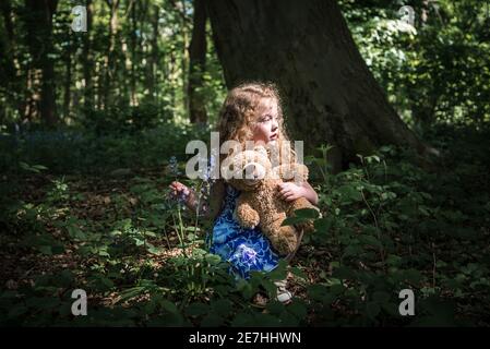 Belle jeune fille long cheveux bouclés dans la robe bleue éclairée par le soleil tenant bluebell et ours en peluche dans la sombre eerie vieille forêt créepy seule Banque D'Images