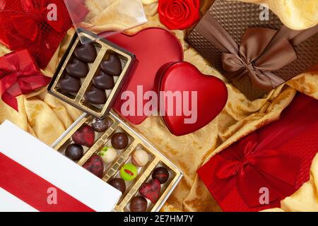 Arrière-plan de la Saint-Valentin, composition de paquets-cadeaux chocolats et boîtes en forme de métal coeur rouge, avec noeuds et rubans, vue du dessus sur tissu doré. Banque D'Images