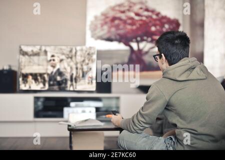 Garçon avec des lunettes un pull à capuche gris regardant la télévision avec le peinture de l'arbre rouge en arrière-plan Banque D'Images