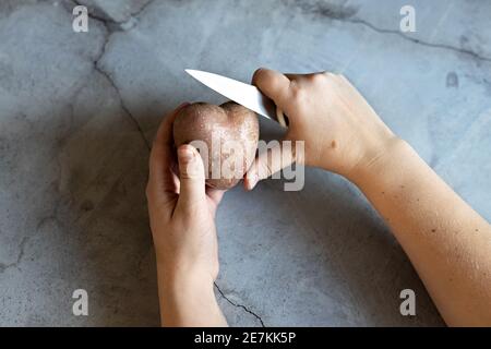 Pommes de terre en forme de coeur et un couteau de cuisine aux mains des femmes sur le fond d'une table en marbre gris. Cuisson des aliments, nettoyage des légumes. Vue depuis un Banque D'Images