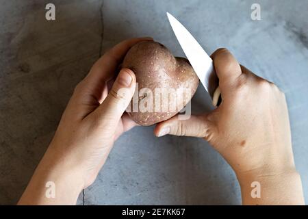 Pommes de terre en forme de coeur et un couteau de cuisine aux mains des femmes sur le fond d'une table en marbre gris. Cuisson des aliments, nettoyage des légumes. Vue depuis un Banque D'Images