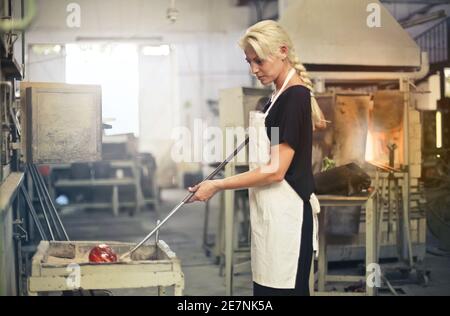 Femme blonde façonnant le verre chaud avec des outils dans le usine de verre Banque D'Images