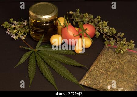 Une feuille de cannabis, un pot avec de l'huile de cbd, des tomates et des herbes aromatiques sur fond noir. L'image illustre la relation entre l'alimentation et le cannabis Banque D'Images