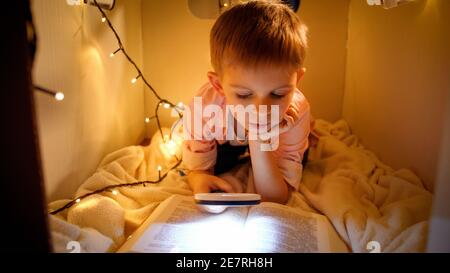 Portrait d'un petit garçon intelligent qui lit un livre de grandes histoires la nuit. Enfant jouant dans la maison en carton de jouet. Concept de l'éducation et de la lecture des enfants dans l'obscurité Banque D'Images