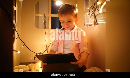 Portrait d'un garçon heureux msiling utilisant un ordinateur tablette et de naviguer sur Internet tout en jouant dans sa maison en carton jouet ou tente la nuit. Concept de l'enfant Banque D'Images