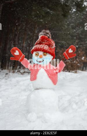 bonhomme de neige heureux en bonnet tricoté rouge, chandail, mitaines, masque médical avec sourire peint sur elle se dresse dans la forêt enneigée. Vie intéressante pendant COVID 19 PAN Banque D'Images