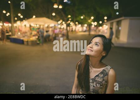 Young happy Asian woman smiling et penser contre le marché de nuit de Hua Hin en Thaïlande avec des gens l'achat et la vente de divers biens Banque D'Images