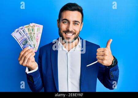 Un jeune homme d'affaires hispanique portant un costume d'affaires portant des billets de roupie indienne souriant heureux et positif, faisant le pouce vers le haut excellent et signe d'approbation Banque D'Images