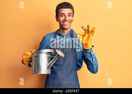 Jeune homme amérindien africain portant un tablier de jardinier et des gants tenant l'arrosage peut faire signe ok avec les doigts, souriant sympathique gesturant excellent sym Banque D'Images