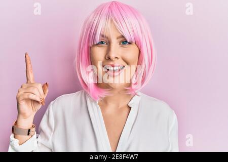 Jeune femme blonde portant une perruque rose pointant du doigt vers le haut avec une idée réussie. Sortie et heureuse. Numéro un. Banque D'Images