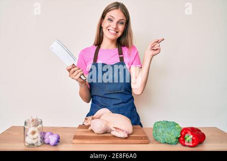 Jeune fille blonde portant un tablier de cuisinier cuisiner poulet sourire heureux en pointant avec la main et le doigt sur le côté Banque D'Images