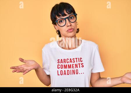Belle femme brunettte portant des commentaires sarcastiques chargement de t-shirt expression sans indice et confuse avec les bras et les mains soulevées. Doute concept. Banque D'Images